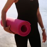 Woman holds a pink yoga mat by the water.
